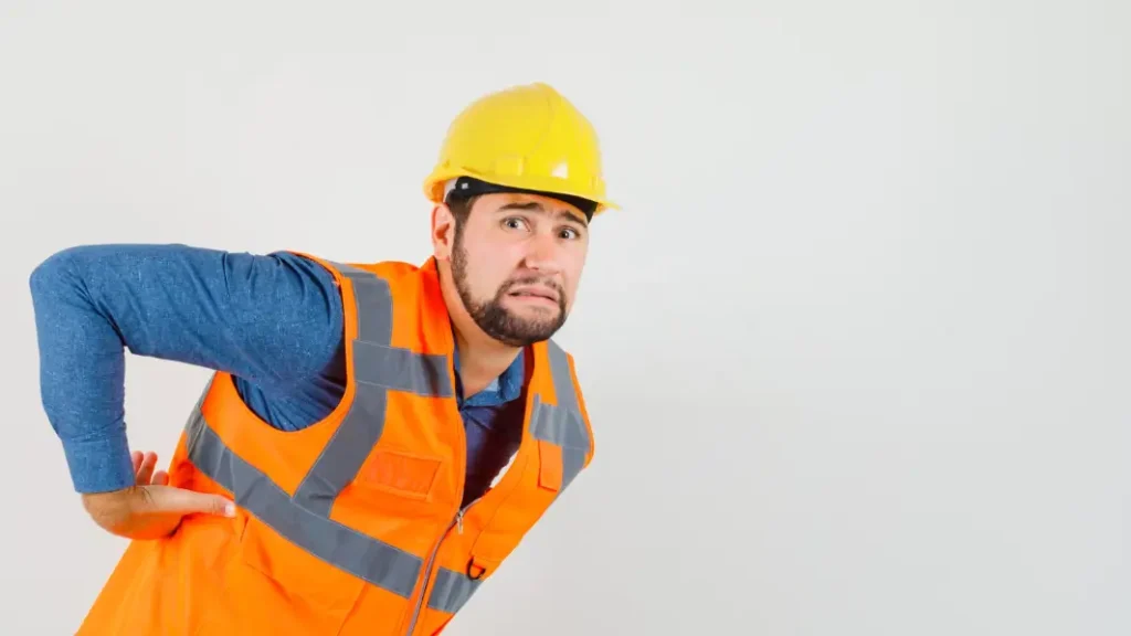 Trabajador de la construcción con casco y chaleco de alta visibilidad sujetándose la zona lumbar con expresión de dolor.
